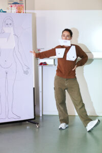 Hey Körper- Szenenbild - Eine Frau hat aus Papier ausgeschnittene Brüste und einen Mund angeklebt. Neben ihr sieht man die Lebensgroße Zeichnung einer Frau, von der die Körperteile ausgeschnitten sind. 